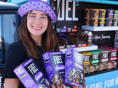 A smiling woman shows off Fuel10K's products, stood by an open Piaggio Ape.