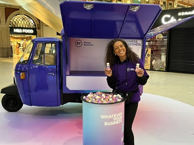 A female staff member smiles in front of the BT Piaggio Ape, holding samples.