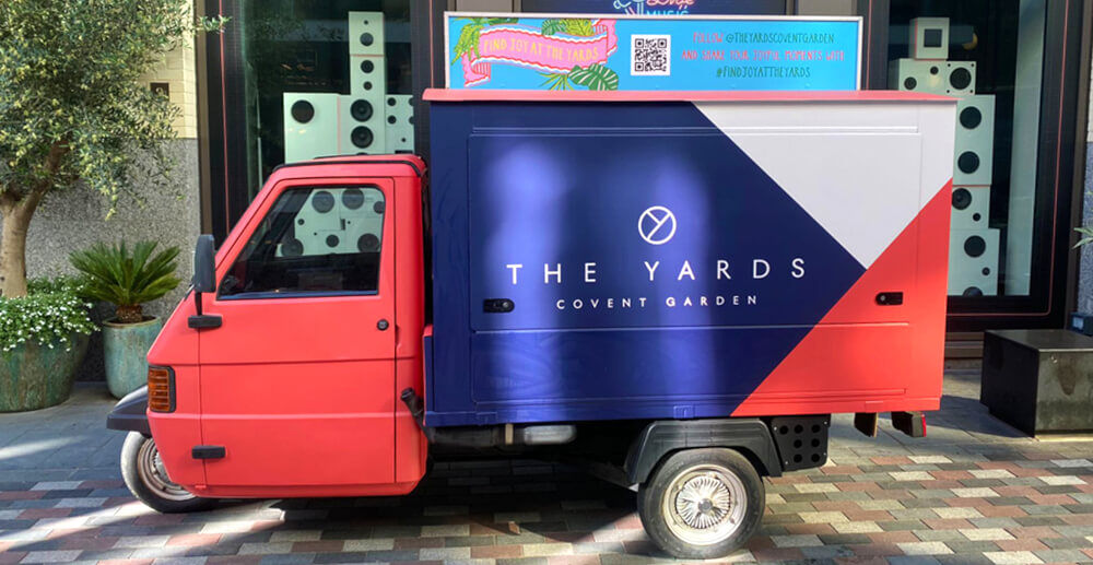 The Piaggio Ape 50, branded for The Yards and viewed from the side, with a red wrap.